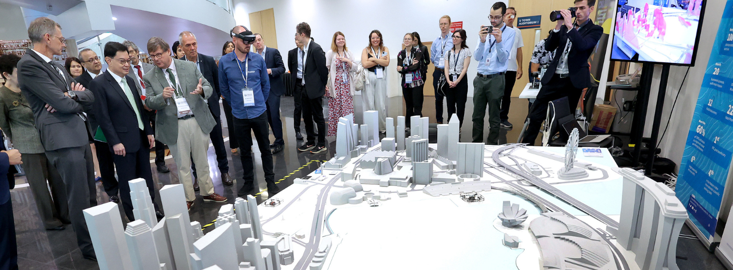 People viewing a scale model of a city with skyscrapers, a harbor, and roadways. One person wears a VR headset.